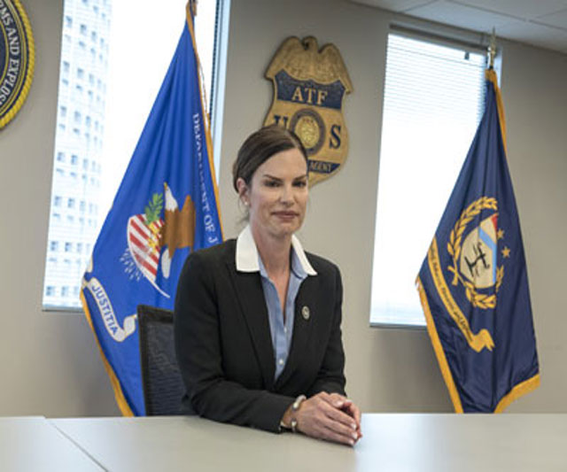 Area Supervisor Mary Harmon Eliason seated in between the DOJ and ATF flag, in front of the ATF seal and ATF badge replica.