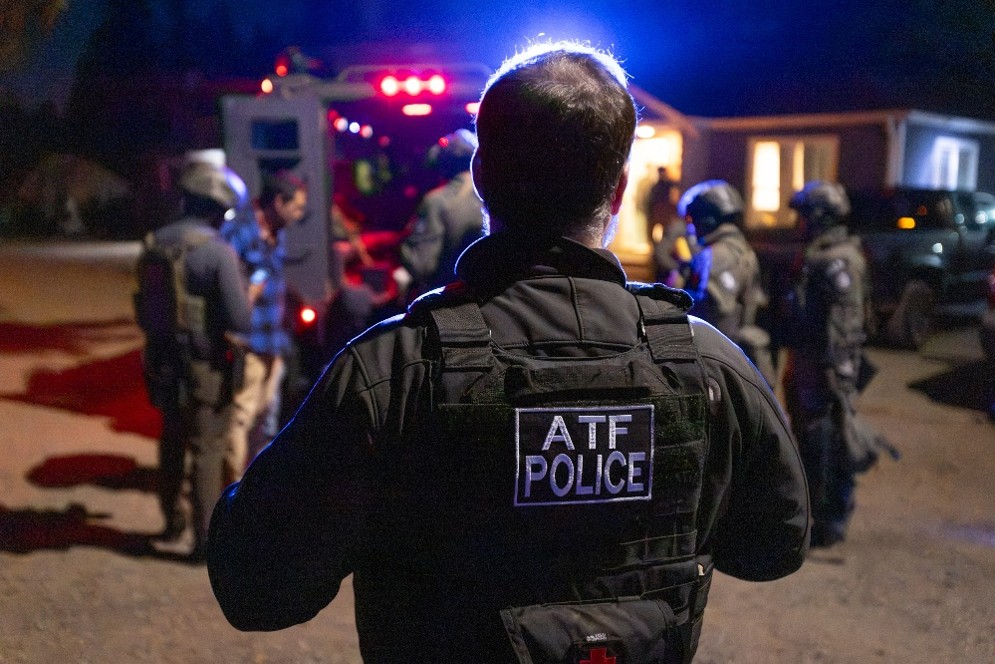 A person wearing a vest with the lettering "ATF Police" stands by multiple law enforcement personnel and a large vehicle.