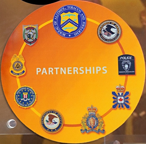 Image of the seals and badges of the ATF, INS, Charlotte-Mecklenburg Police, Canadian Security Intelligence Service, Royal Canadian Mounted Police, DOJ, FBI, Diplomatic Security, and Iredell County Sheriff, who were all involved in the investigation.