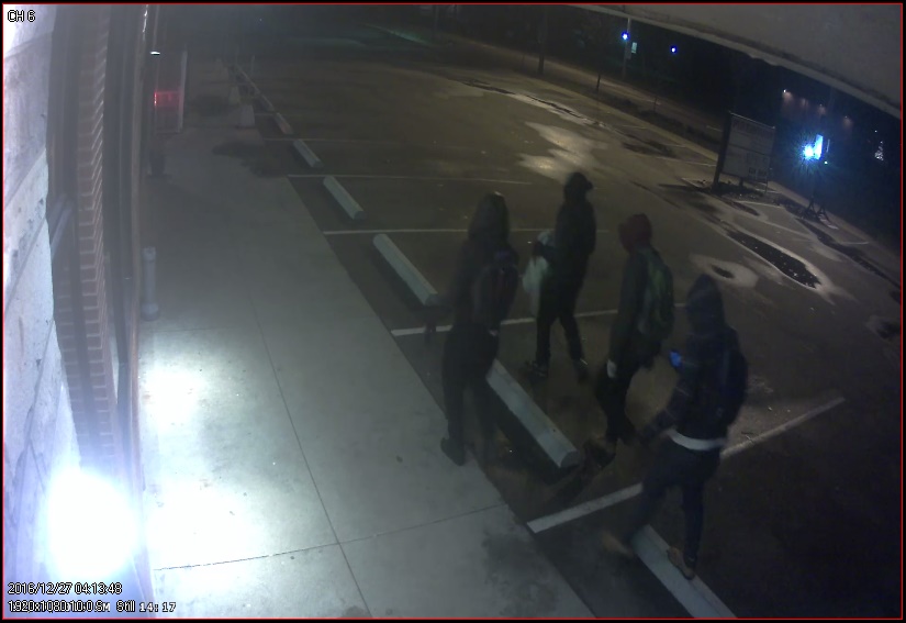 Image of four people wear jackets with the hoods over their heads walking through a parking lot. Three of the four are also wearing backpacks. 