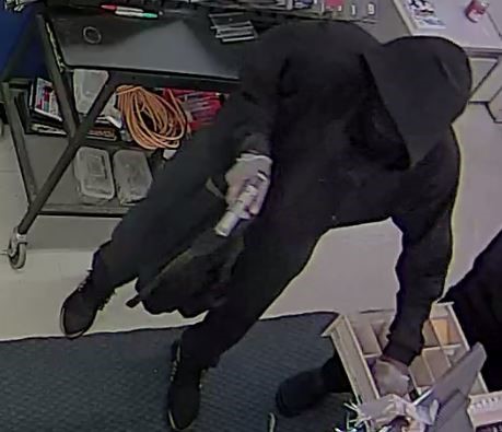 Image of suspect wearing black clothing, holding a firearm in one hand and removing money out of the register with the other. 