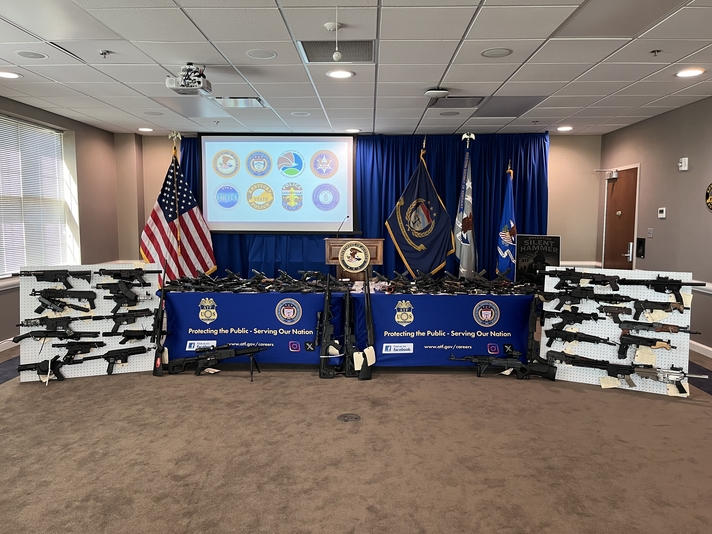 A photograph of a table displaying numerous firearms alongside various law enforcement seals and flags.