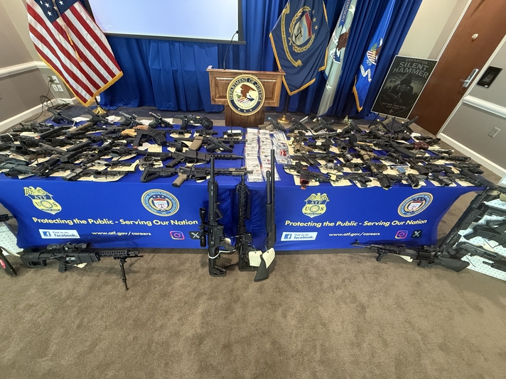 An up-close photograph of a table displaying numerous firearms alongside various law enforcement seals and flags.