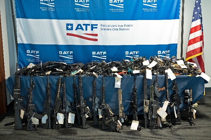 Large number of firearms displayed on and leaning against a table.