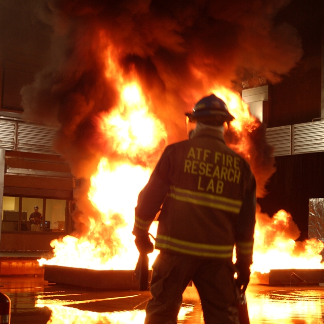 An individual in an ATF Fire Research Lab jacket in front of a fire